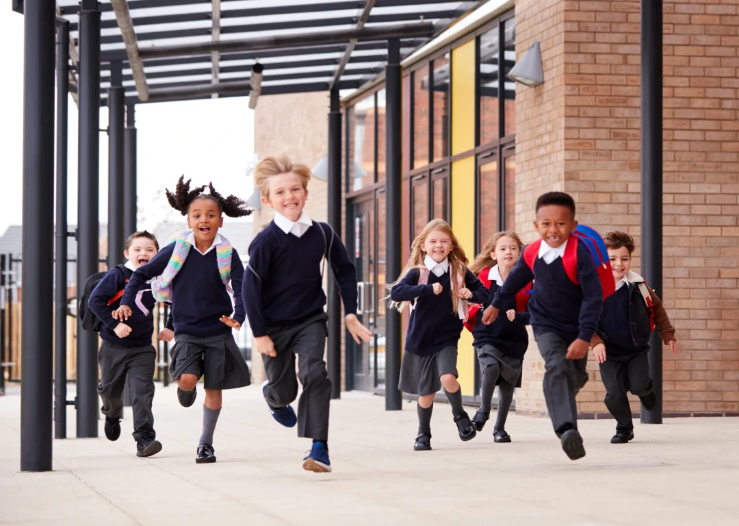 Primary school children in uniforms and wearing backpacks running next to school.