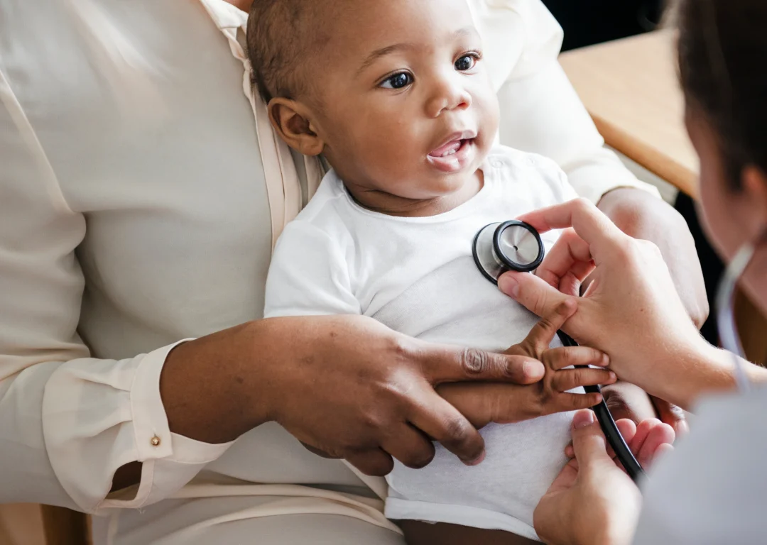Baby is sat on mother's lap while doctor uses stethoscope.