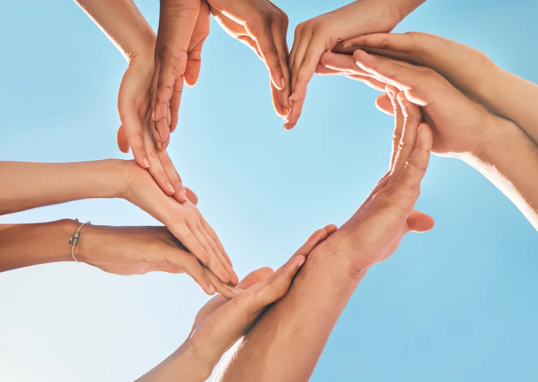 Hands of all ages together forming the shape of a heart against a clear blue sky, representing heart screening and heart health.