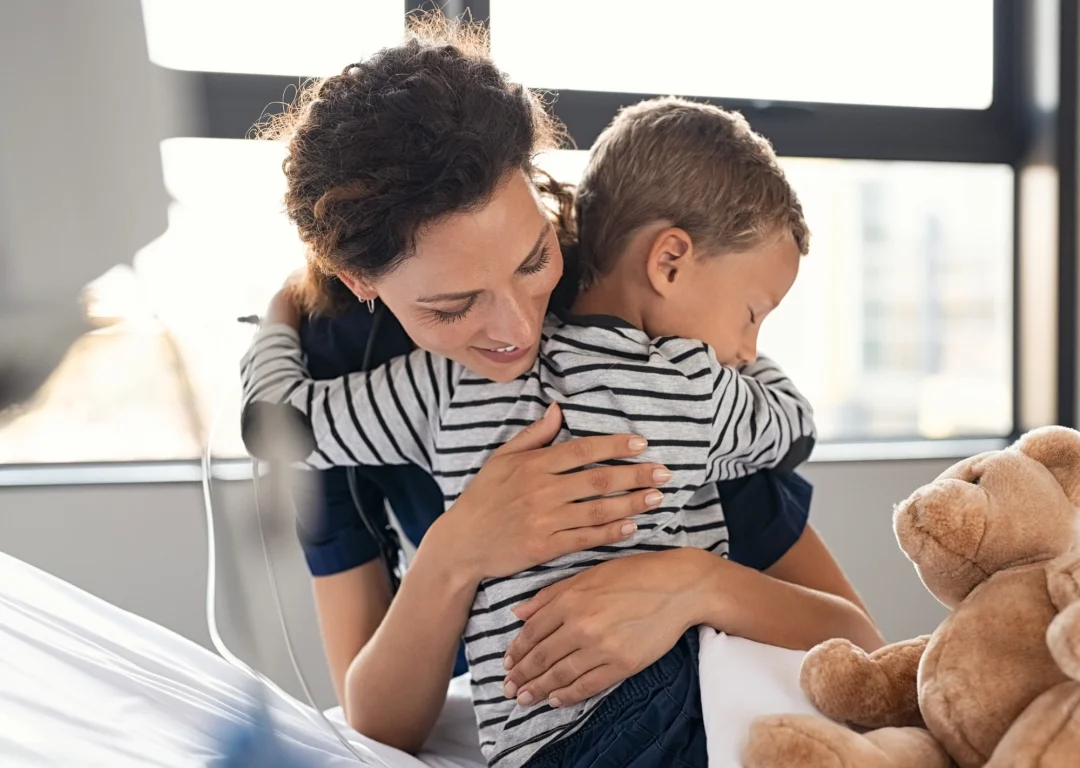 Young boy in hospital bed with teddy bear being embraced by his mother.